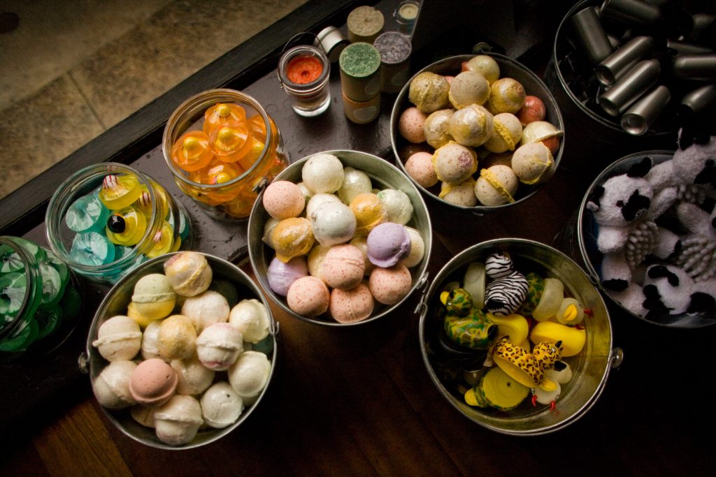 A display of various bath bombs in metal buckets and glass jars, with additional colorful rubber ducks and plastic containers around them on a wooden surface.