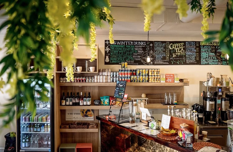 Coffee shop interior with a cozy atmosphere, featuring a menu board, counter with various drinks and snacks, overhead greenery, and refrigerated display cases.