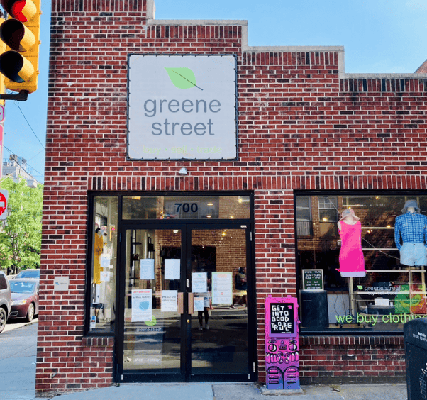 Brick storefront of Greene Street clothing store with large sign, glass entrance, and visible window displays. Bright pink clothing donation bin labeled Get Good Stuff, Do Good Stuff is outside.