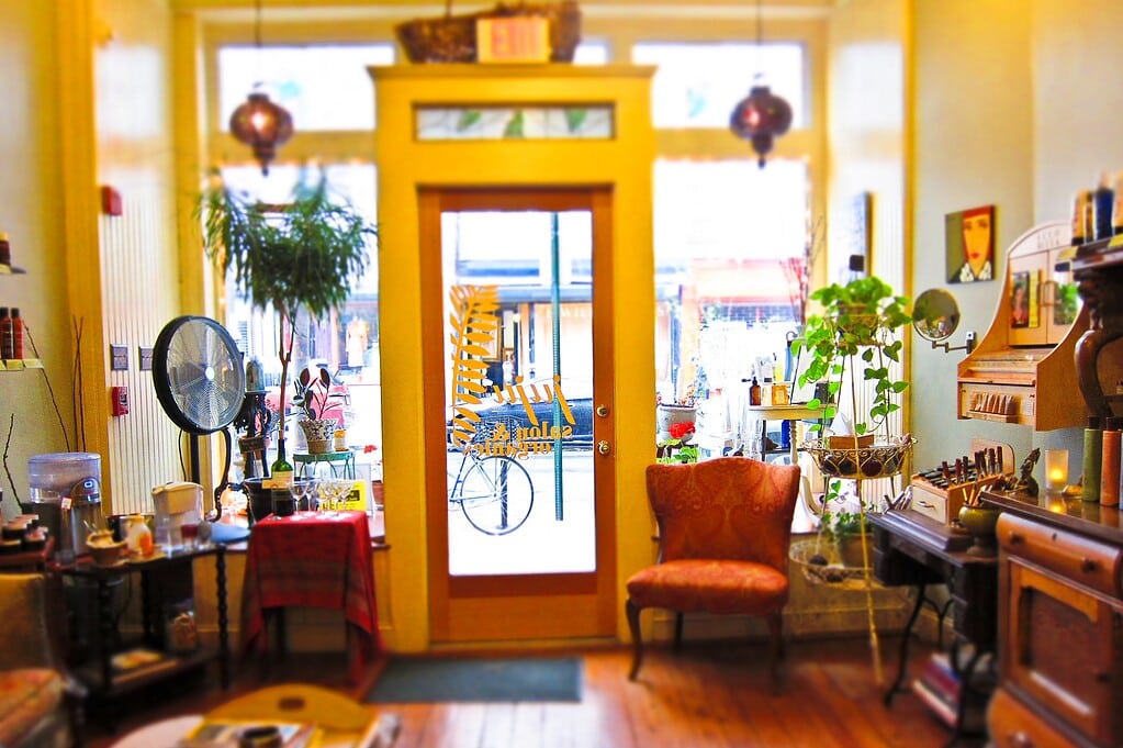 Cozy shop interior with vintage decor, wooden furniture, and plants. Large windowed door opens to a street view. A bicycle and people are visible outside. Warm lighting enhances the ambiance.