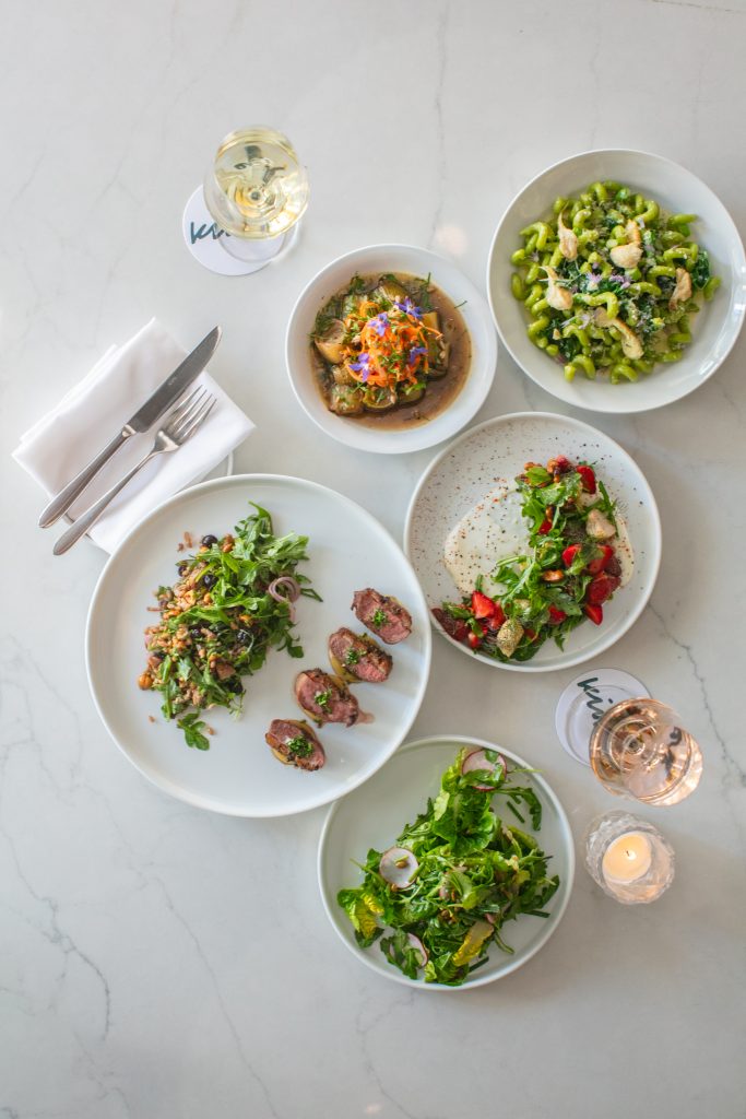 Aerial view of a table with plates of salads, grilled meat, pasta, and a glass of white wine on a marble surface.