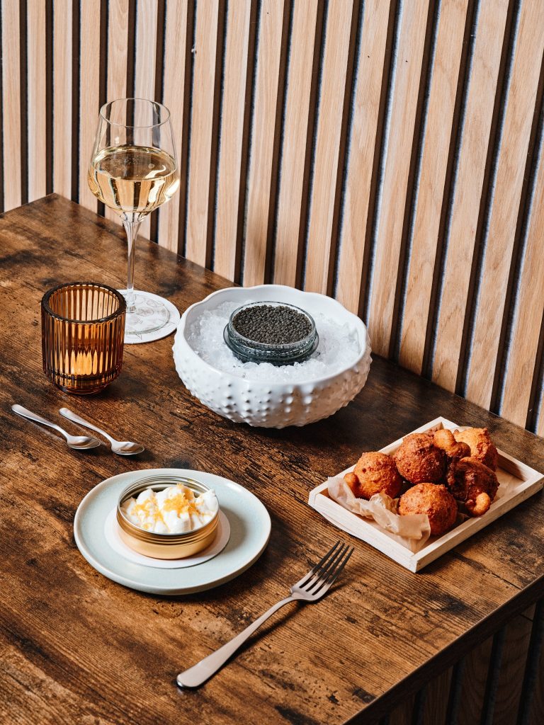 A table setting with a glass of white wine, a small dish of caviar in a white bowl, a plate with butter, a tray of bread pieces, a candle, and cutlery on a wooden surface.