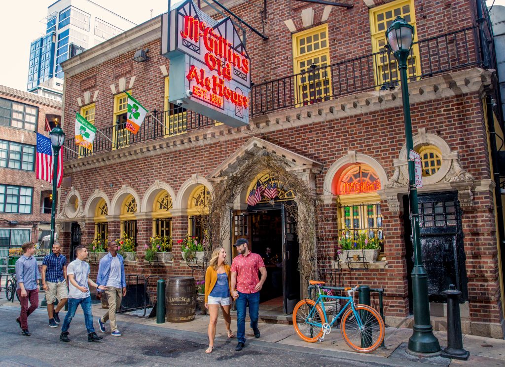 People walking on a city street in front of a brick alehouse with signs. There are two flags, a bicycle, and a lamppost. The building has arched windows and ivy around the entrance.