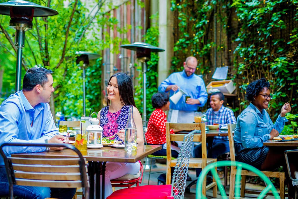 People dining at outdoor tables in a green, leafy setting. Some are eating, some holding drinks. A waiter in the background carries a tray.