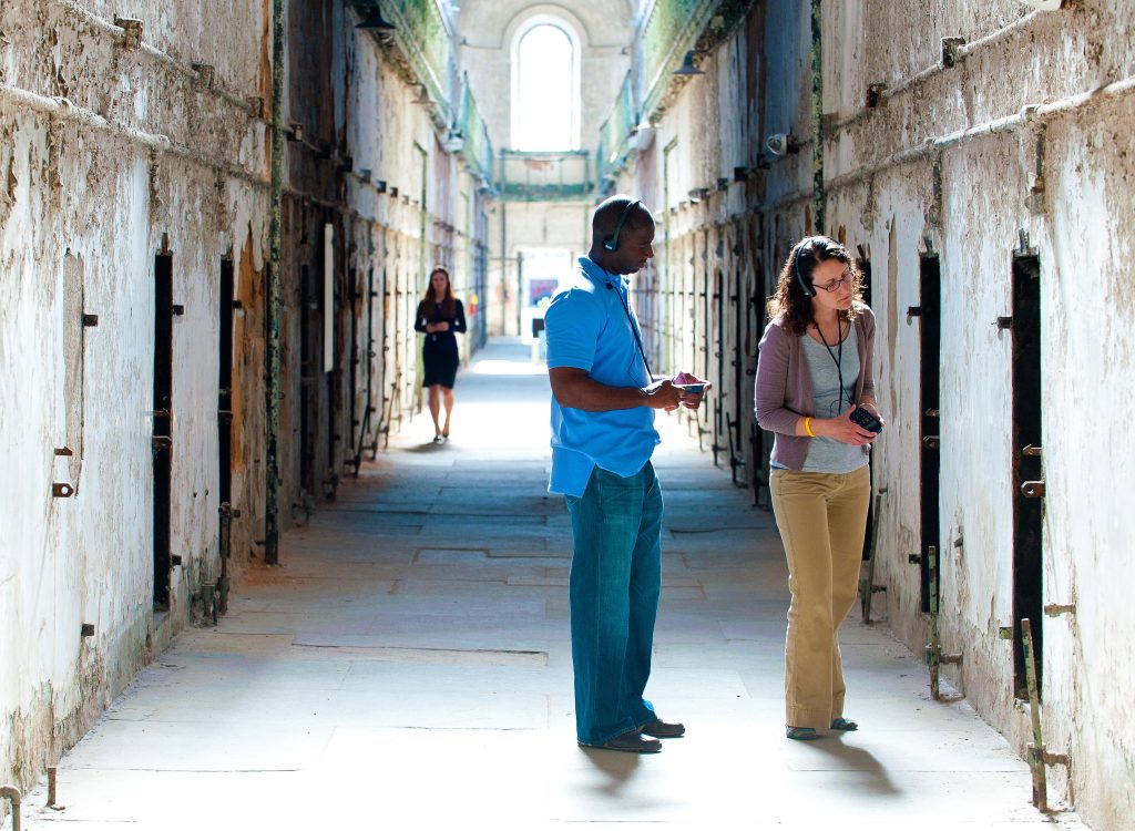 Three people tour an old, sunlit prison cellblock; two examine a cell with audio guides while one walks in the background.