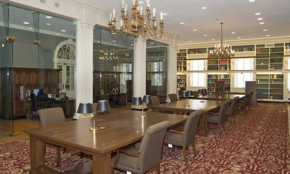 A spacious library reading room with wooden tables, upholstered chairs, chandeliers, bookshelves, and glass partitioned offices in the background.
