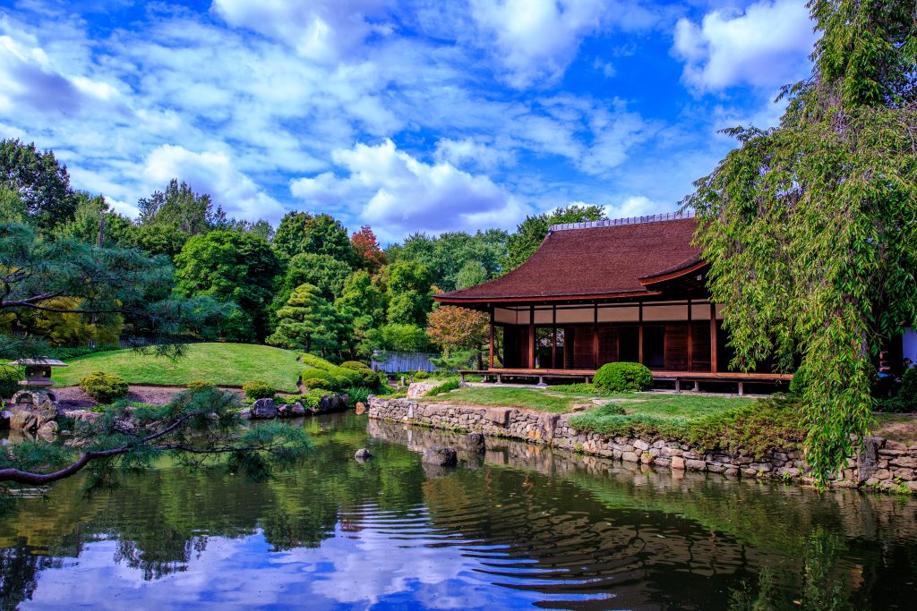 A traditional wooden building stands by a reflective pond surrounded by lush greenery and trees under a blue sky with scattered clouds.