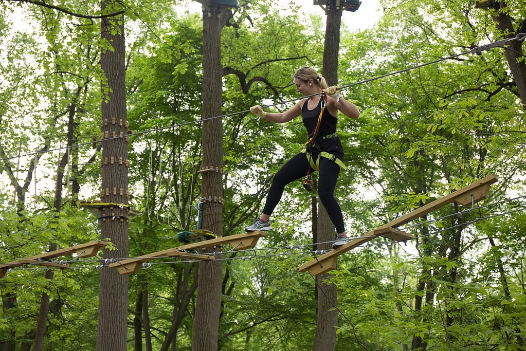 A person wearing safety gear crosses a suspended wooden bridge in a ropes course set among tall trees in a forest.