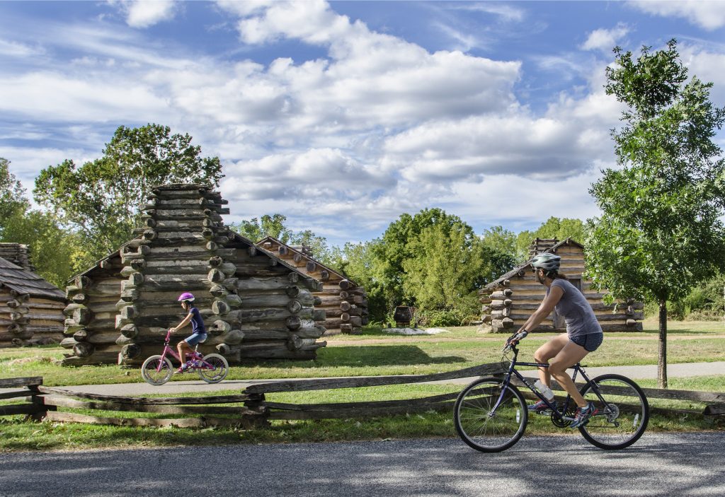 Two people ride bicycles past log cabins on a sunny day, with trees and a partly cloudy sky in the background.