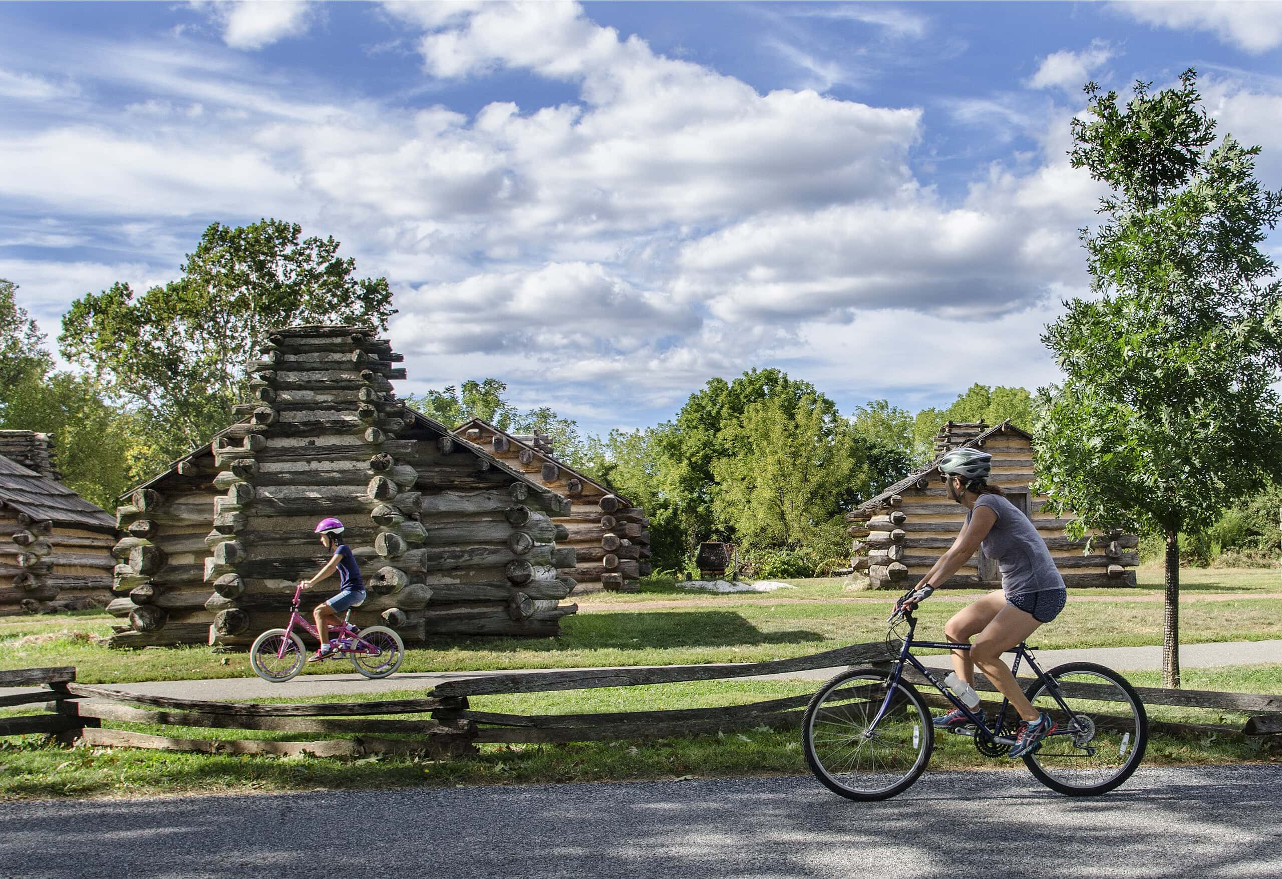 Two people ride bicycles past log cabins on a sunny day, with trees and a partly cloudy sky in the background.