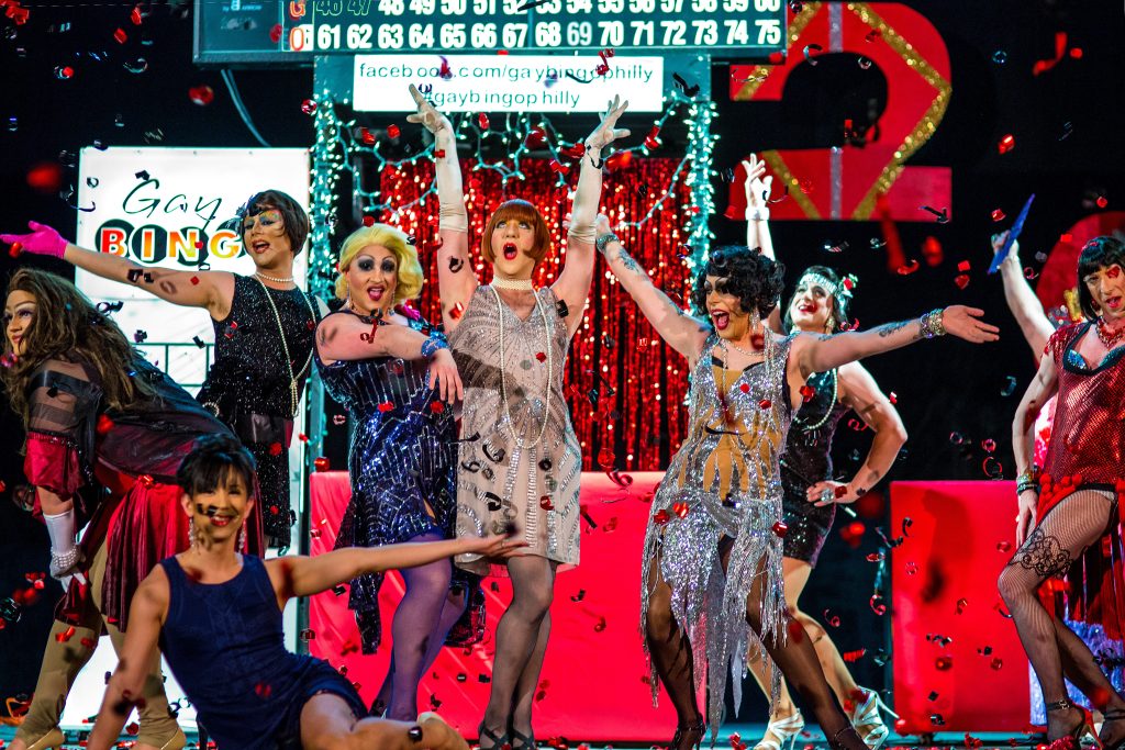 A group of drag performers in colorful costumes dance and pose on stage, surrounded by confetti, with bingo signage and decorations in the background.