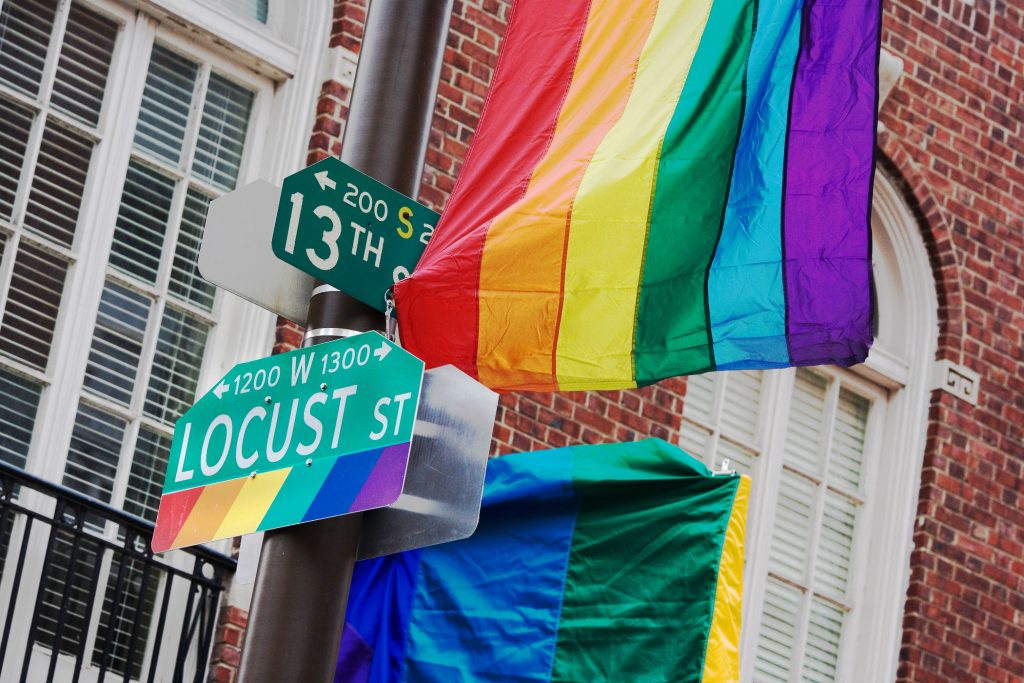 Street signs for 13th Street and Locust Street are decorated with rainbow colors, with large rainbow flags hanging nearby in front of a brick building.