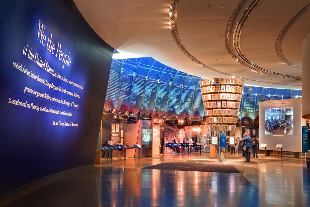 Exhibit hall at the National Constitution Center with a large “We the People” wall, a cylindrical photo display, and people viewing interactive exhibits.