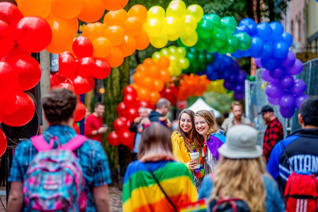 People walk under rainbow balloon arches at an outdoor event. Two women in the center, draped in rainbow flags, smile and pose for a photo. Many attendees wear colorful outfits.