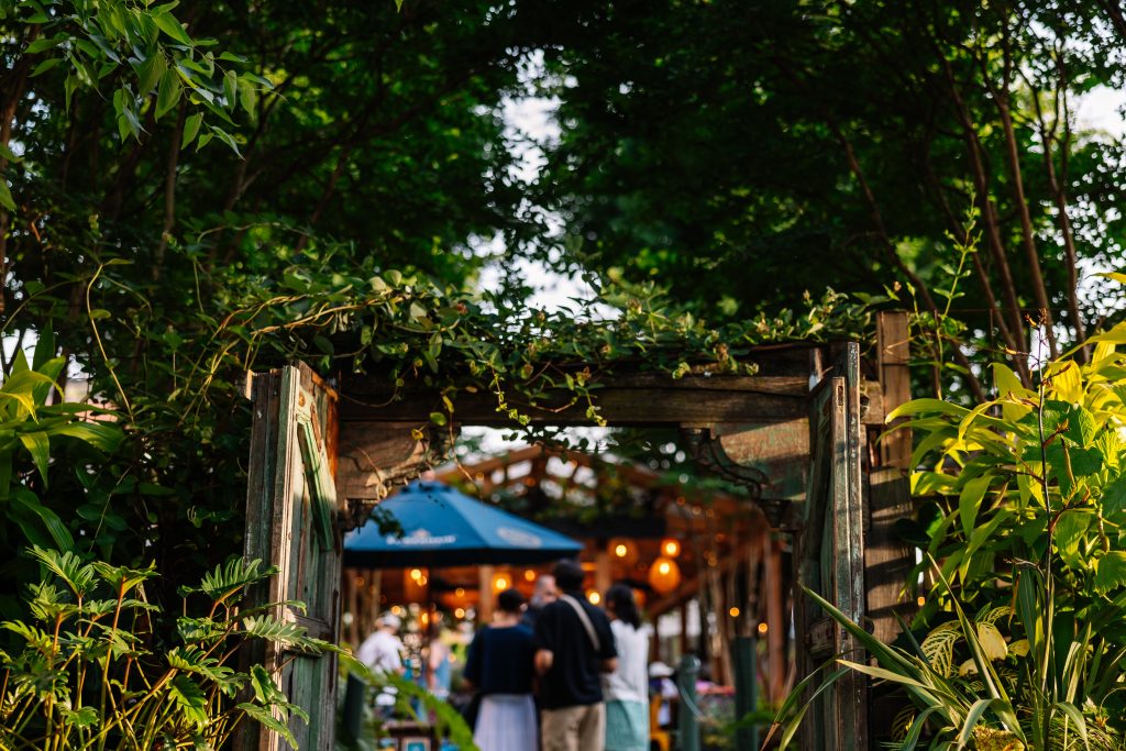 An open wooden garden gate surrounded by greenery leads to a group of people gathered under string lights and a blue umbrella in the background.
