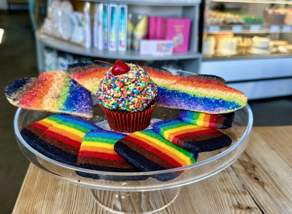 A glass cake stand with rainbow heart cookies, rectangular rainbow cookies, and a cupcake topped with rainbow sprinkles and a cherry, displayed in a bakery.