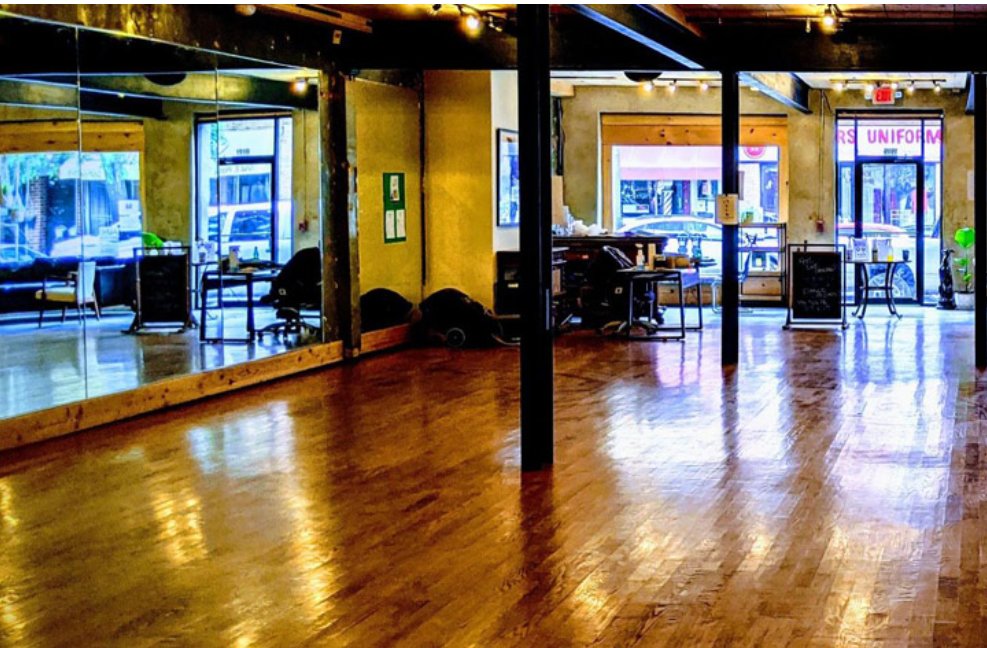 A spacious dance studio with wooden floors, large mirrors on one wall, desks and chairs by the windows, and natural light coming through the front windows.